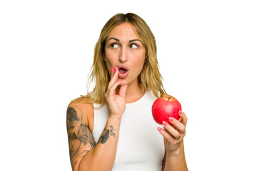 Young caucasian woman holding an apple isolated on green chroma background is saying a secret hot braking news and looking aside