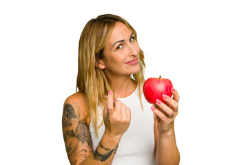 Young caucasian woman holding an apple isolated on green chroma background pointing with finger at you as if inviting come closer.