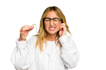 Young otorhinolaryngologist caucasian woman holding hearing aid isolated on green chroma background covering ears with hands.