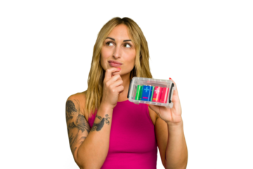 Young caucasian woman holding battery box isolated on green chroma background looking sideways with doubtful and skeptical expression.