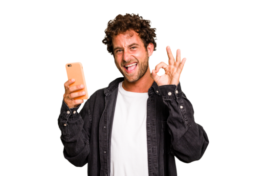Young caucasian man using mobile phone isolated cheerful and confident showing ok gesture.