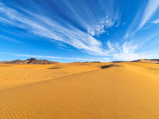 Fototapeta na wymiar Textures sand ripples in foreground and a rocky mountain with a blue sky and stratus clouds with motion effect. Colorful smartphone photography in Tadrart Rouge, Djanet Algeria. Tassili N'Ajjer Sahara