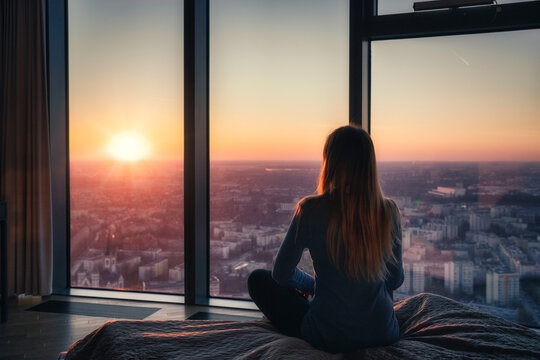 A young woman sitting on a bed and watching the sunset through the windows from a high floor, luxurious apartment with a view on the city.