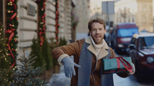 Caucasian Crazy Man Going Dancing On The Decorated Street Listening To Music Using Earphones Singing Holding The Christmas Gift. People And Christmas Holidays Concept