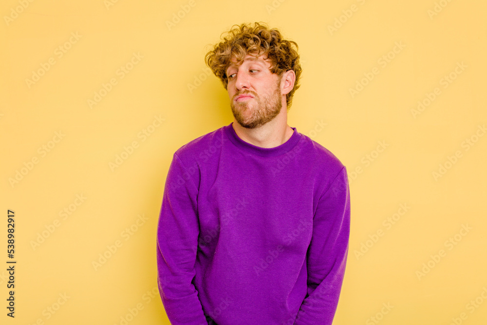 Wall mural Young caucasian man isolated on yellow background shrugs shoulders and open eyes confused.