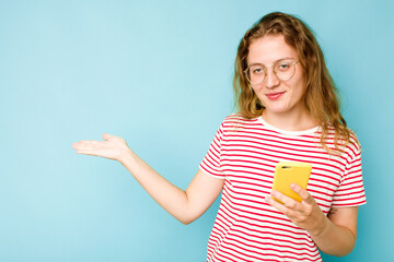 Young caucasian woman holding mobile phone isolated on blue background showing a copy space on a palm and holding another hand on waist.