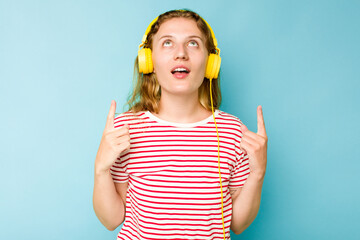 Young caucasian woman wearing headphones isolated on blue background pointing upside with opened mouth.