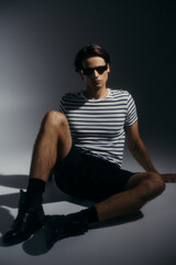 Fototapeta premium Trendy model in sunglasses and shoes posing on grey background with lighting and shadow