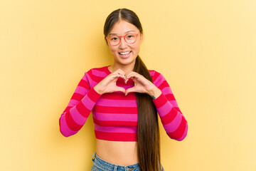 Young Asian woman isolated on yellow background smiling and showing a heart shape with hands.