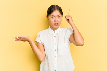 Young Asian woman isolated on yellow background holding and showing a product on hand.