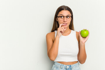 Young Indian woman holding an apple isolated on white background relaxed thinking about something looking at a copy space.