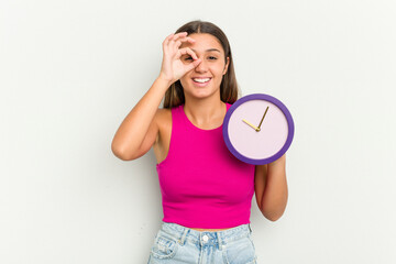 Young Indian woman holding a clock isolated on white background excited keeping ok gesture on eye.