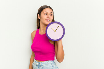 Young Indian woman holding a clock isolated on white background looks aside smiling, cheerful and pleasant.