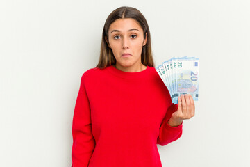 Young Indian woman holding a banknotes isolated on white background shrugs shoulders and open eyes confused.