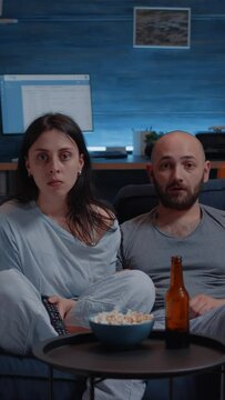 Vertical video: Shocked astonished young couple looking at documentary show at tv having surprised facial expression, eating popcorn sitting on sofa. Concentrated adults watching tv late night