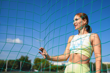Women and sport. Beautiful girl in summer sportswear stands behind the sports net and smiles on the green grass of a stadium. Middle aged sportswoman