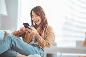 Fototapeta na wymiar Happy young asian woman browsing surfing wireless internet on mobile phone while sitting a couch in living room at home, Shopping online via website