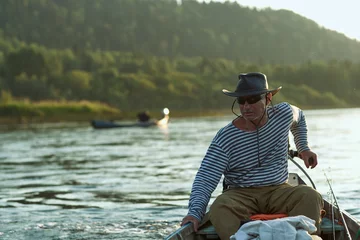 Foto op Canvas A brutal man, a fisherman aged 60+ in a vest and a black hat, drives a motor boat on the river. Sunset time on the river. The concept of tourism and recreation. High resolution photo. Horizontal frame © Marina