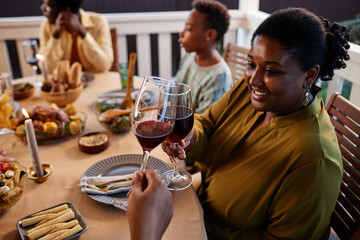 Side view portrait of smiling black woman clinking wine glasses at dinner table outdoors in cozy setting