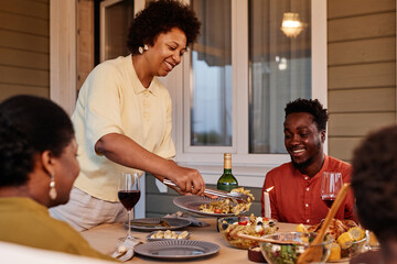 Portrait of smiling African American woman serving food to family while enjoying dinner together outdoors in evening