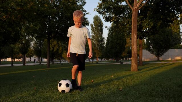 A little athletic boy trains with a soccer ball in the city park on the green grass in summer at sunset.