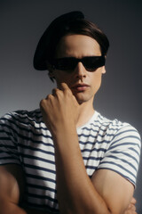 Brunette man in sunglasses and striped t-shirt touching chin isolated on grey with lighting