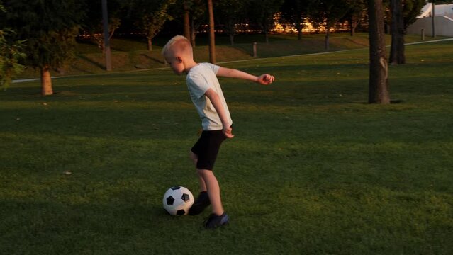 A little boy trains on a green lawn with a soccer ball in a city park in the summer at sunset.