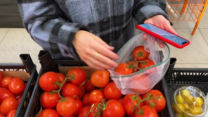 Close up of female hands choosing tomatoes at the supermarket. girl putting vegetables into reusable produce eco bag. female customer noting the purchase in shopping list on smartphone.