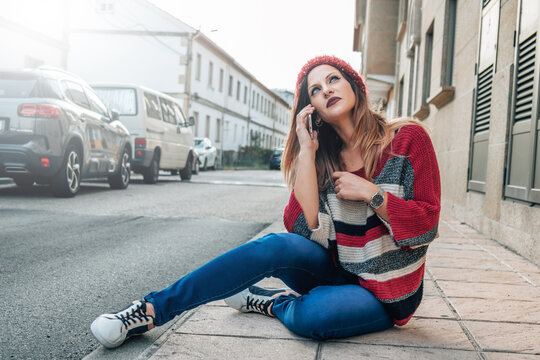 young woman with hat and sweater sitting on the street talking on a mobile phone