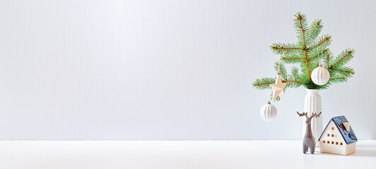 Obraz na płótnie Canvas Christmas, New Year home decor. Empty white wall mock up with green fir branches in a vase, christmas little house and deer on a white table. Mock up for displaying works