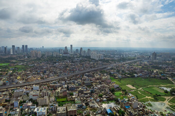 aerial drone shot over gurgaon showing monsoon clouds with light rays falling on ground crowded with homes houses, sohna highway feilds and water pools around under construction buildings