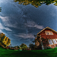 Milky Way above the Swedish traditional wooden red house at the left side of night scene in garden. Starry sky, green grass.