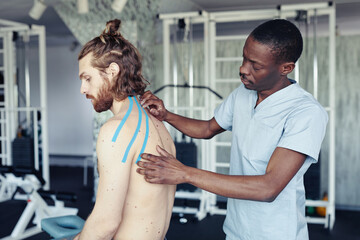 African young therapist applying tapes on back of patient to treat ligaments before sport training