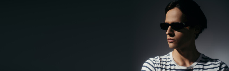Brunette man in sunglasses and striped t-shirt standing isolated on grey with lighting, banner
