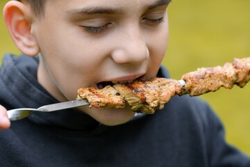 The boy eats fried meat on a fire strung on skewers.