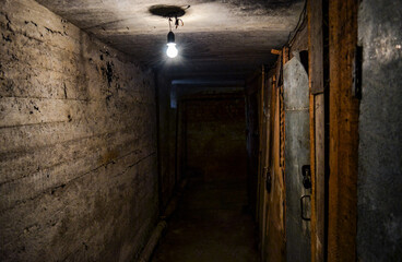 A scary dark concrete corridor in the basement, lit by a single light bulb hanging from the low ceiling. Copy space