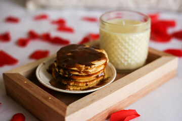 plate with pancakes and sauce next to an aromatic candle on a wooden tray on a bed with petals