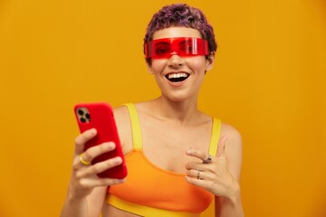 Woman blogger surprise in unusual millennial glasses taking selfies on her phone sporting brightly colored clothes against an orange studio background, free space