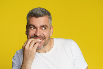 Scared of competitor handsome middle aged man bitting his fingernails. Freelancer in stress of project deadline. Shocked freelancer man eating fingers in white t-shirt isolated on yellow background