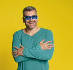 Fashionable handsome middle aged man posing with arms folded wearing casual and blue glasses isolated on yellow background. Attractive man posing in long sleeve t-shirt. Grey haired mature man