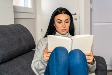 Young woman with long dark hair lying on the sofa while reading a book