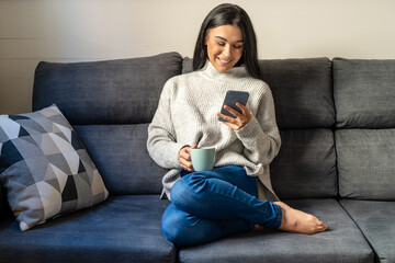 Obraz premium Young woman with long dark hair sitting on the sofa by a window looking at the phone with a cup of coffee or tea in the other hand