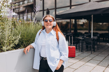 Lovely happy young woman in sunglasess listening to music through wireless earphones on urban background. Music lover enjoying music. Lifestyle concept. Girl walking on city street