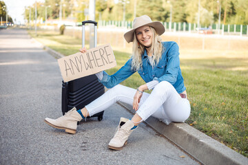 Happy woman wait passing car sitting with suitcase and cardboard poster on roadside of empty road. Blonde lady in hat escape from city by auto stop to go anywhere. Travelling, hitchhiking, vacations