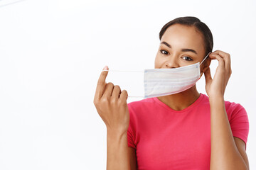 People during pandemic. Smiling black woman puts on medical face mask to protect from covid-19, standing in tshirt over white background