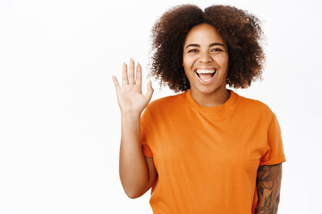 Friendly smiling african american woman saying hello, waving hand at you to greet, hi gesture, standing over white background in orange tshirt © Cookie Studio
