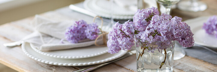 Beautiful table decor for a wedding dinner with a spring blooming lilac flowers. Celebration of a special event. Fancy white plates, and wineglasses. Countryside style. Banner