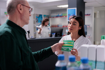 Pharmacy worker showing pills package to senior customer during medical consultation explaining prescription. Elderly man buying supplements, drugs to cure disease. Health care support