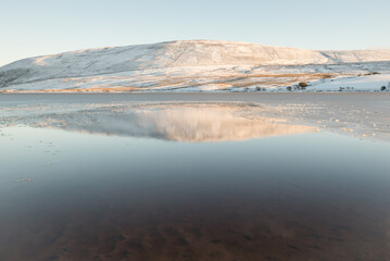 View of a reservoir in the Pentland Hills Regional Park in Edinburgh, Scotland, UK, where a hill covered in snow is reflected on the calm water under a blue sky