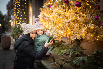 An Asian woman wearing a woolly hat texting her family on the phone with her daughter sat next to an illuminated Christmas decorated tree in the streets of in Edinburgh, Scotland, UK during Christmas